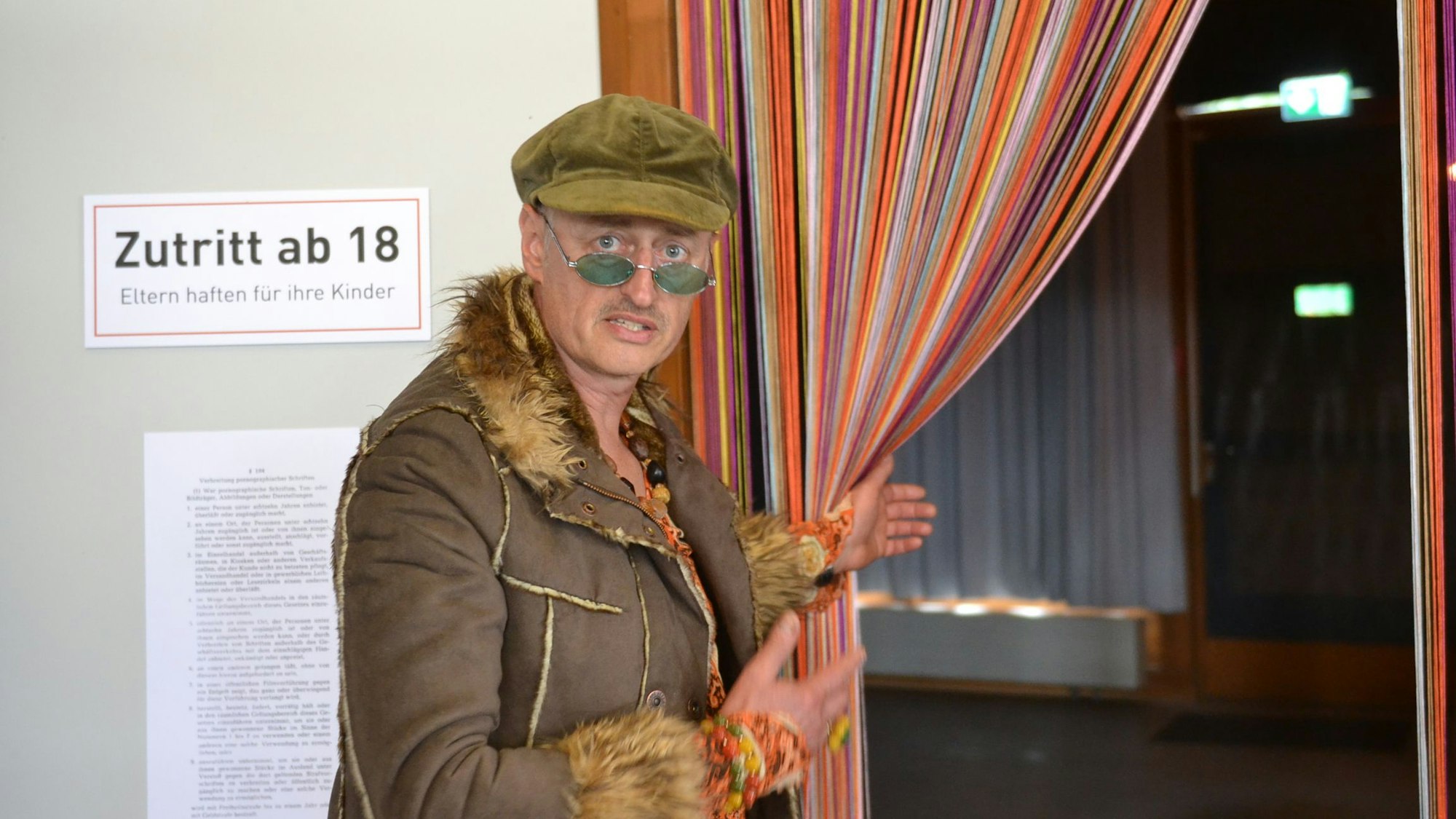 Ein Türsteher hält einen Vorhang auf. Er trägt Schiebermütze, blaugetönte Brille und einen Mantel mit Pelzbesatz. Auf einem Schild steht "Zutritt ab 18".