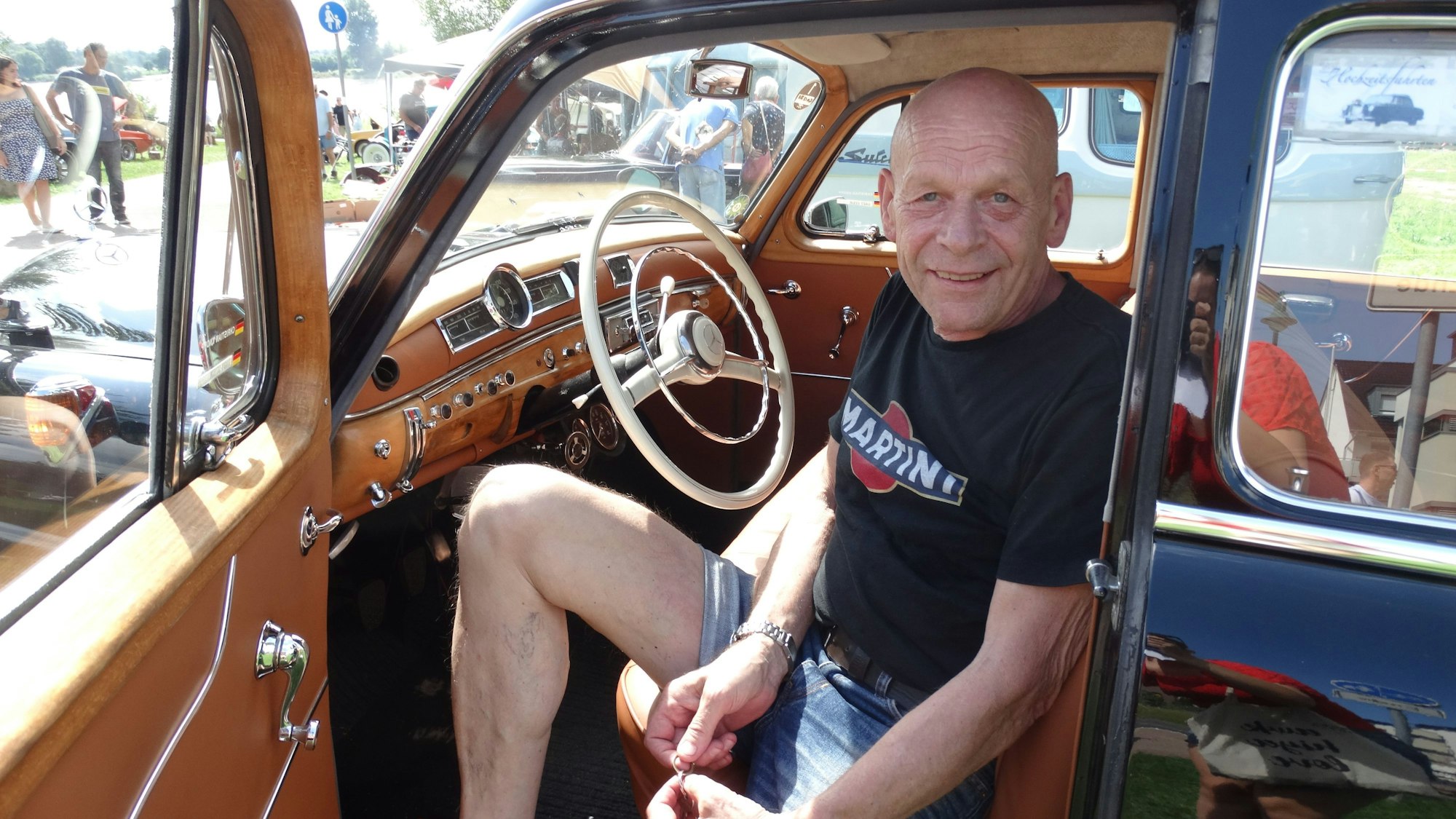 Ein kahlköpfiger Mann in Shorts und T-Shirt sitzt am Steuer eines alten Mercedes mit großem Lenkrad und heller Lederausstattung.