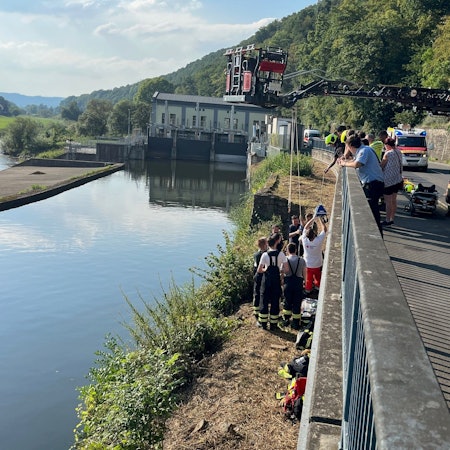An einem breiten Kanal stehen mehrere Rettungskräfte an einer Mauer, die zu einer höher gelegenen Straße gehört. Dort steht die ausgefahrene Drehleiter der Feuerwehr.