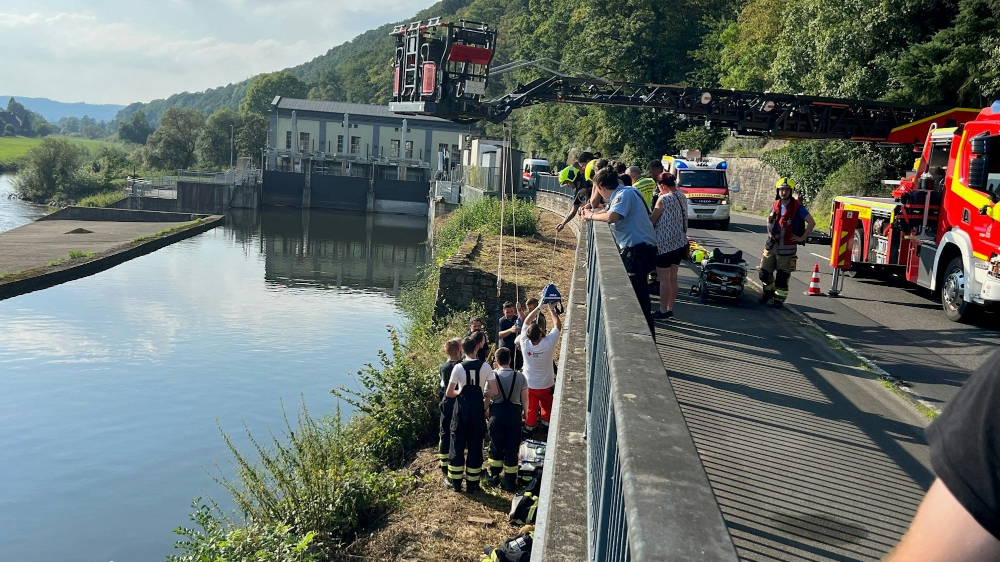 An einem breiten Kanal stehen mehrere Rettungskräfte an einer Mauer, die zu einer höher gelegenen Straße gehört. Dort steht die ausgefahrene Drehleiter der Feuerwehr.