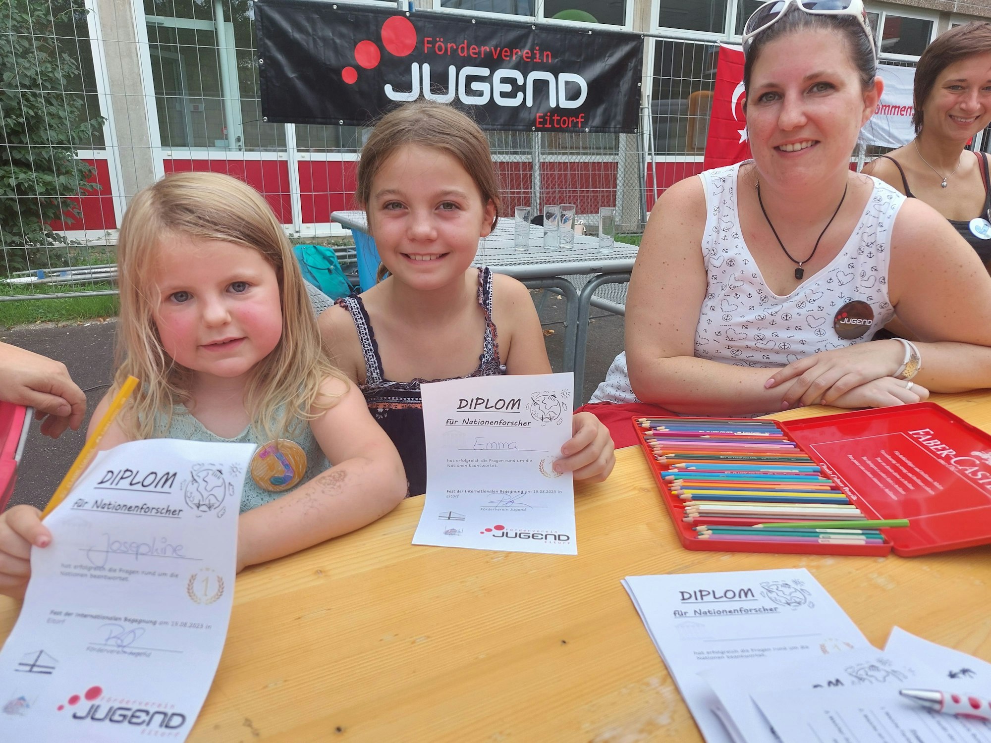 Zwei kleine Mädchen sitzen an einem Tisch und zeigen stolz ein Diplom vor, neben ihnen liegt ein Kasten mit Buntstiften. Eine Frau lächelt in die Kamera.