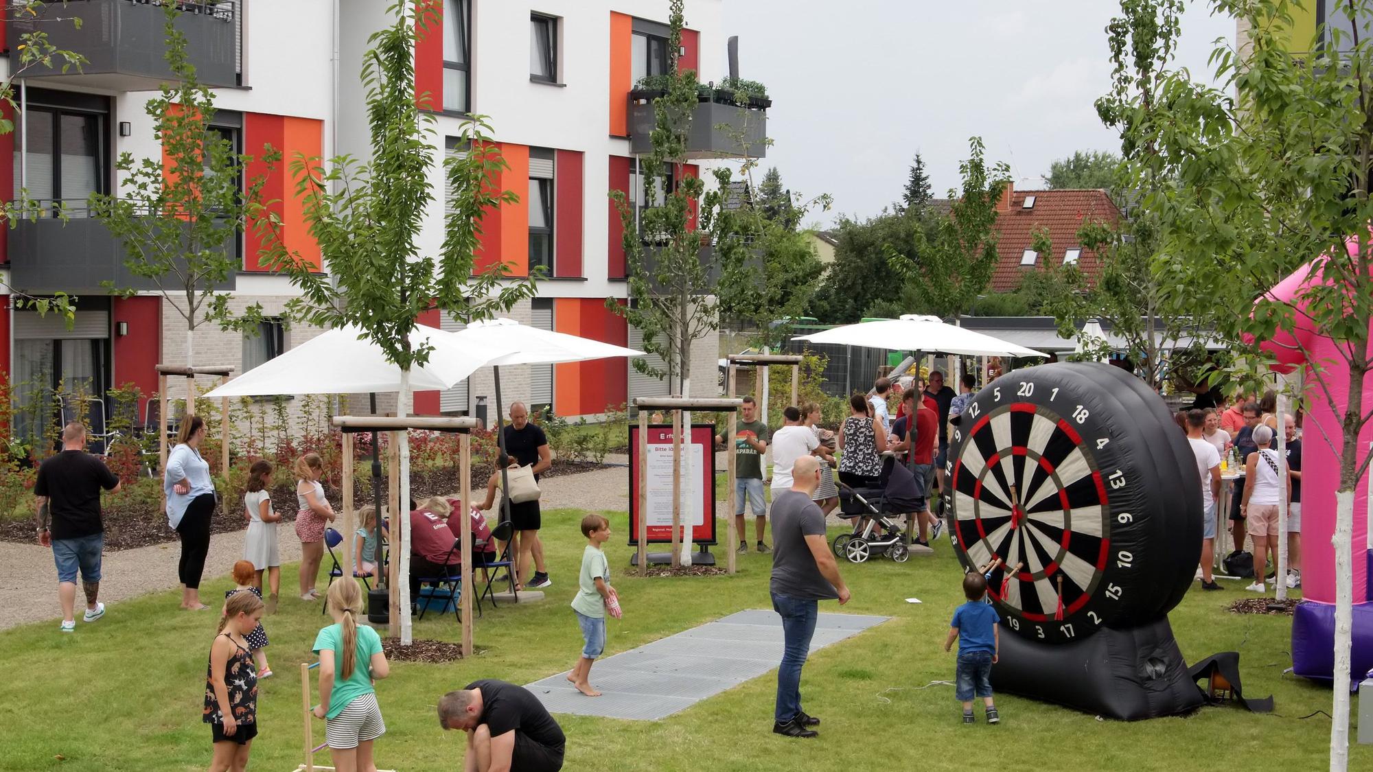 Auf einer WIese mit jungen Bäumen feiern Kinder und Erwachsene mit einer Hüpfburg und einer aufblasbaren Wurfscheibe, im Hintergrund steht ein neugebautes Mehrfamilienhaus.