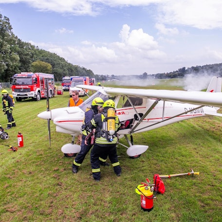 An einem qualmenden Kleinflugzeug stehen Rettungskräfte der Feuerwehr.