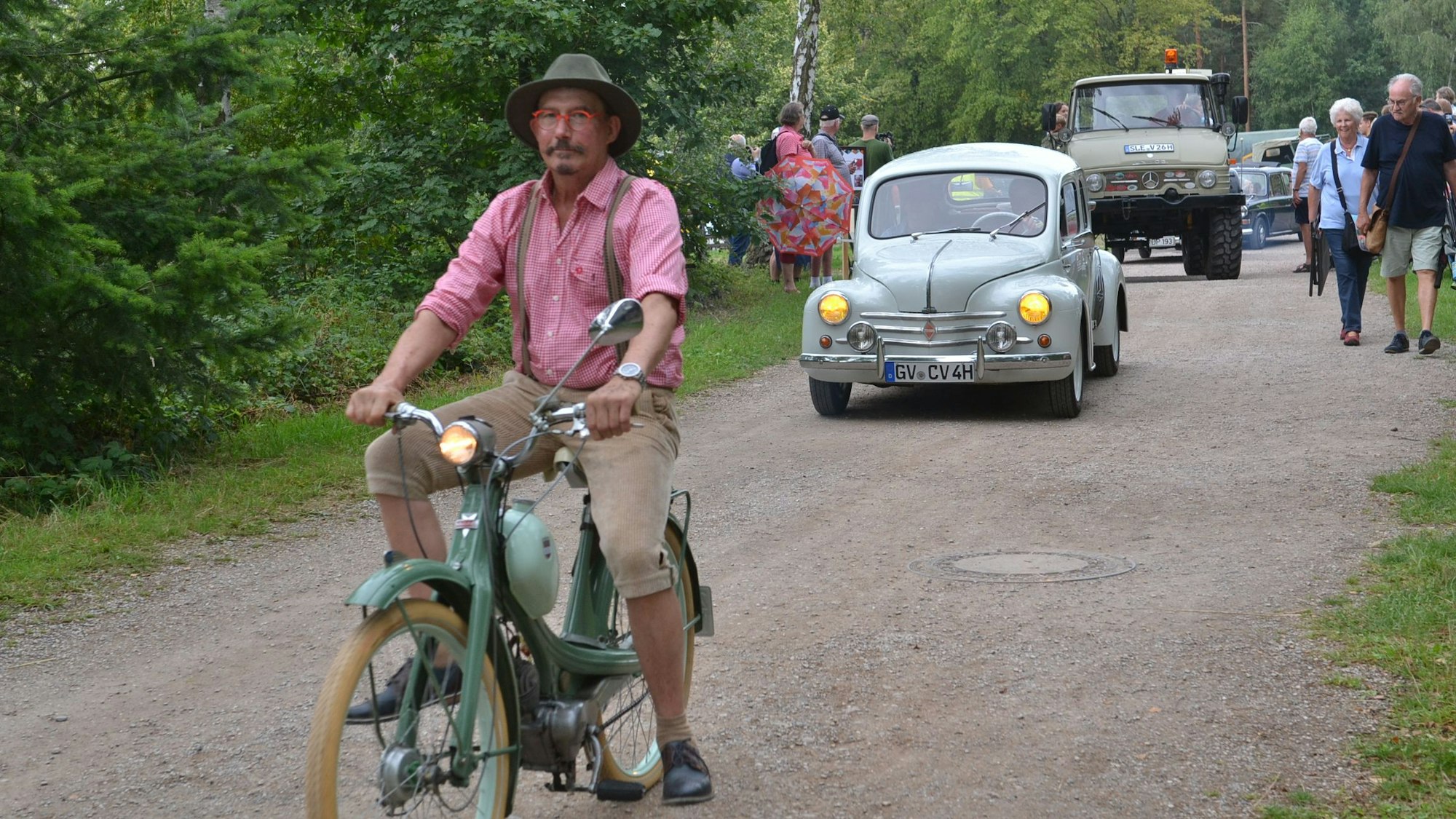 Auf einem Moped sitzt ein Mann. Hinter ihm fahren Oldtimer die unbefestigte Straße entlang.