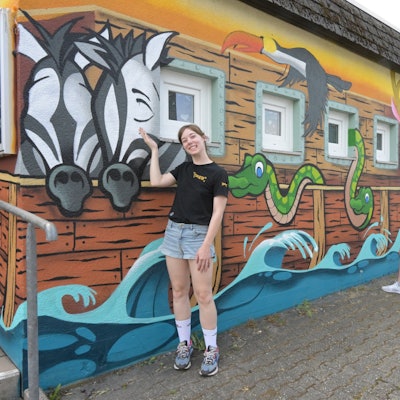 Vor einem Gebäude stehen zwei junge Mädchen und ein Mann. Die Fassade des Hauses wurde mit bunter Farbe bemalt. Das Motiv ist die Arche Noah.