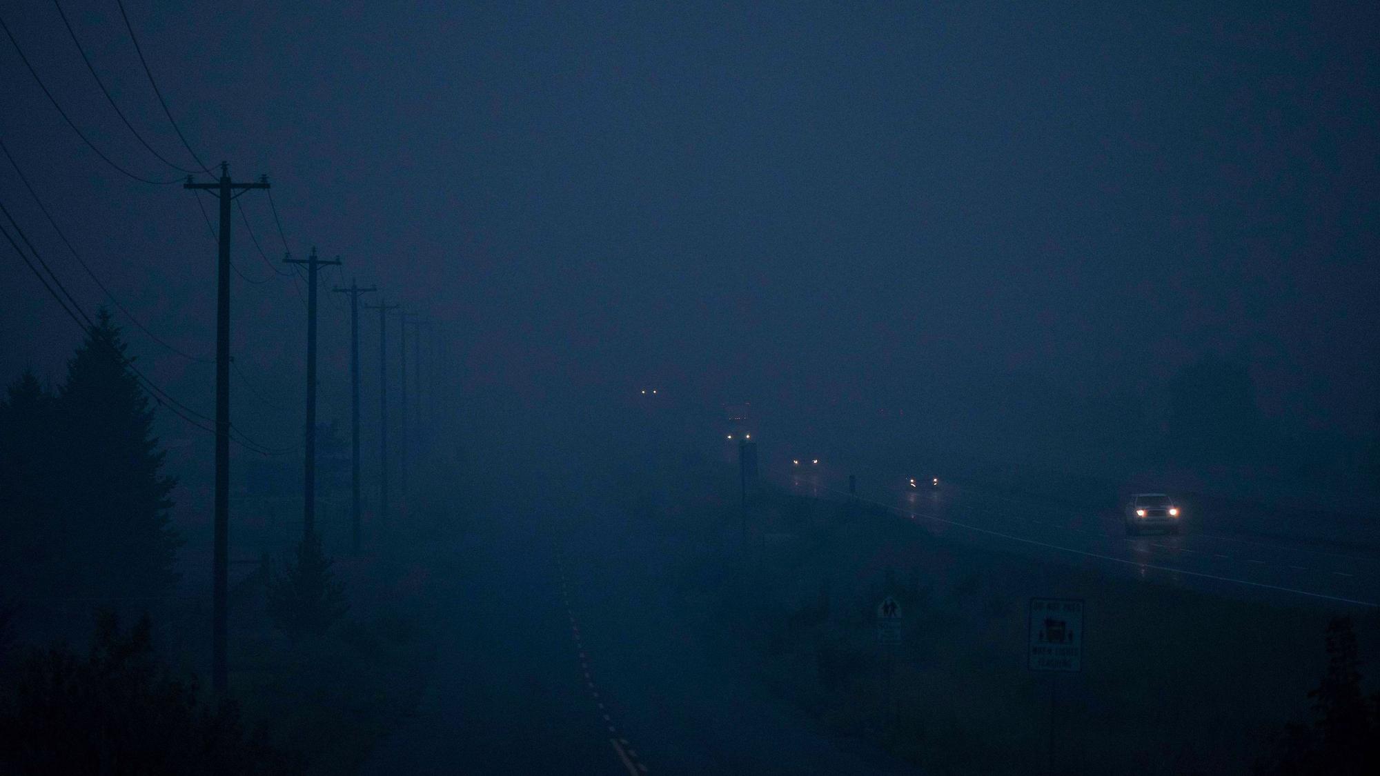 Autos fahren über eine vom Rauch vernebelte Straße, am linken Bildrand sind Silhouetten von Leitungsmasten zu erkennen.