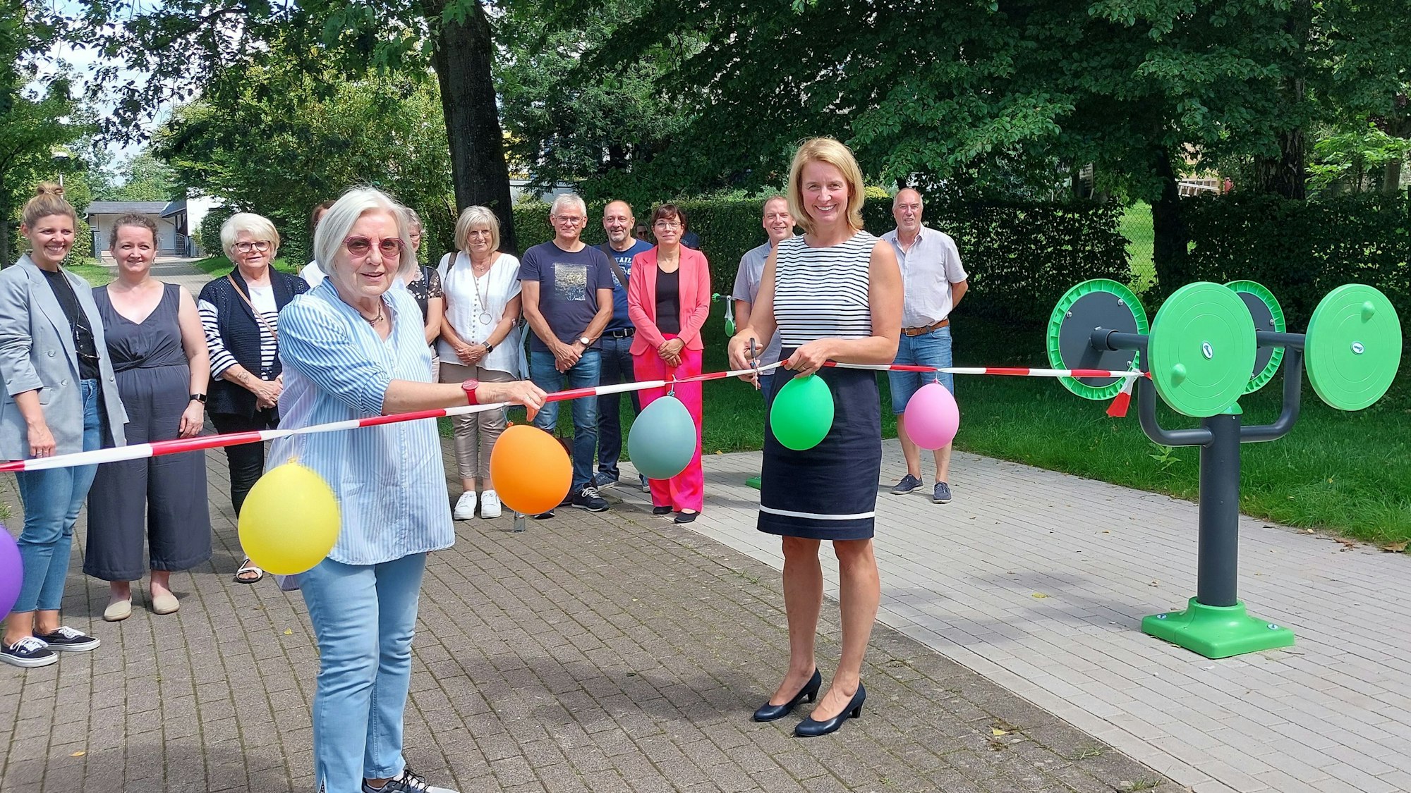 Zwei Frauen schneiden ein Absperrband, geschmückt mit Luftballons durch. Dieses ist an Outdoorfitnessgeräten befestigt.