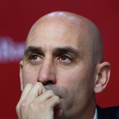 Nahaufnahme von Luis Rubiales Gesicht vor einem unscharfen roten Hintergrund - Rubiales hält eine Hand am Mund und schaut in die Ferne.