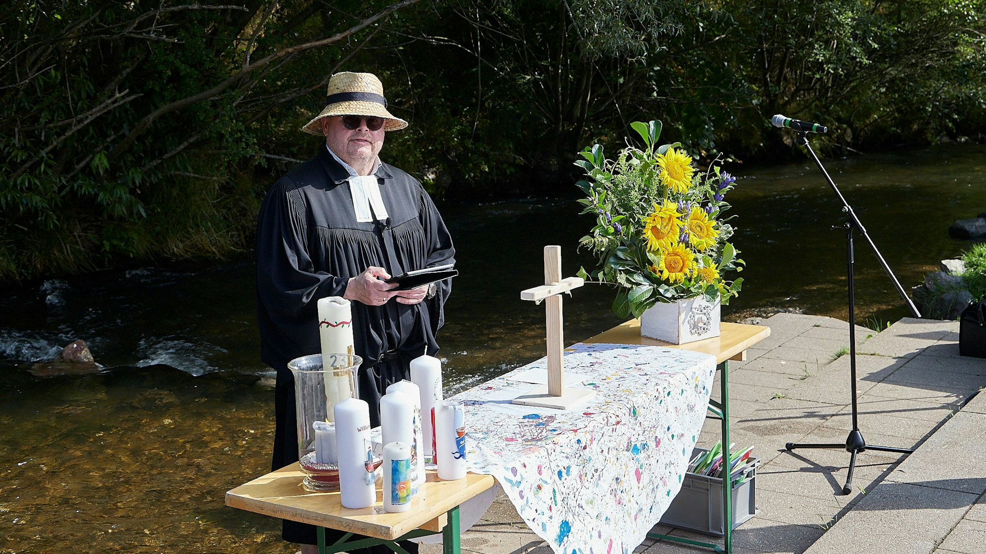 Pfarrer Oliver Joswig trägt Strohhut und Sonnenbrille. Er steht an einem Altar, der   auf einem Holztisch vor der Olef in Hellenthal aufgebaut ist.