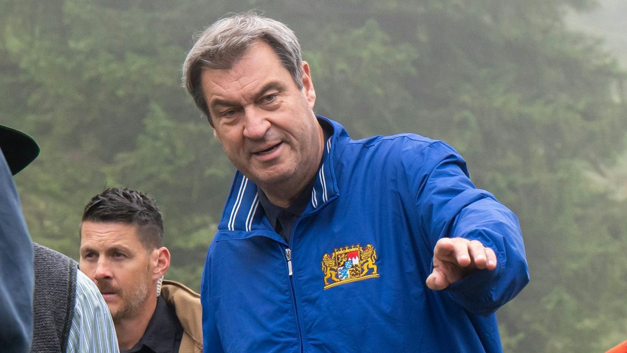 Der bayrische Ministerpräsident Markus Söder (CSU) zeigt bei einer Almumgehung mit diversen weiteren Almbauern in Richtung der Kamera. Er trägt eine blaue Jacke mit dem bayrischen Landeswappen.