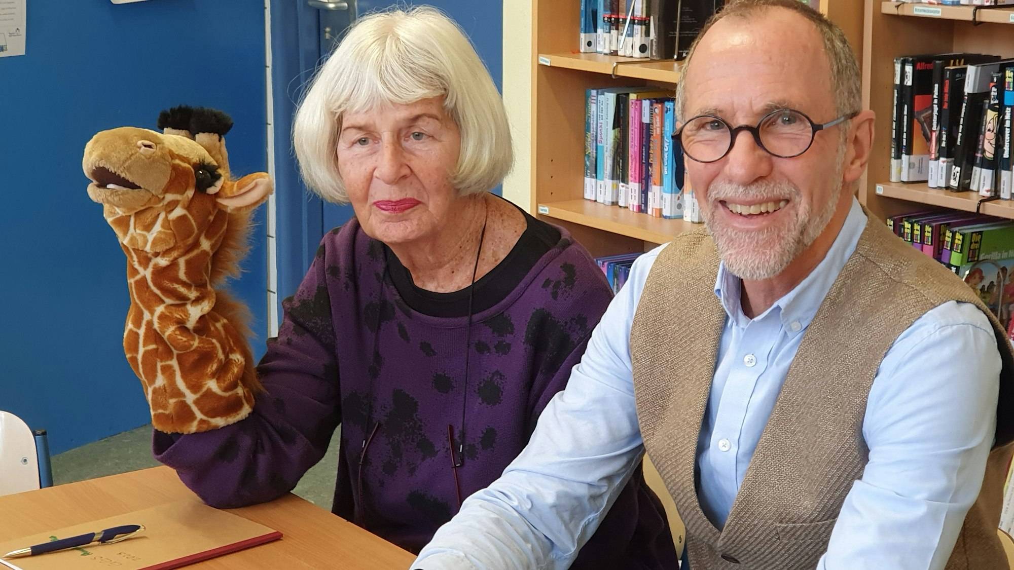 Eine ältere Dame und ein älterer Mann sitzen an einem Pult. Die Senioren hat den Ellenbogen aufgestützt, an ihrem Unterarm steckt eine Giraffen-Handpuppe.