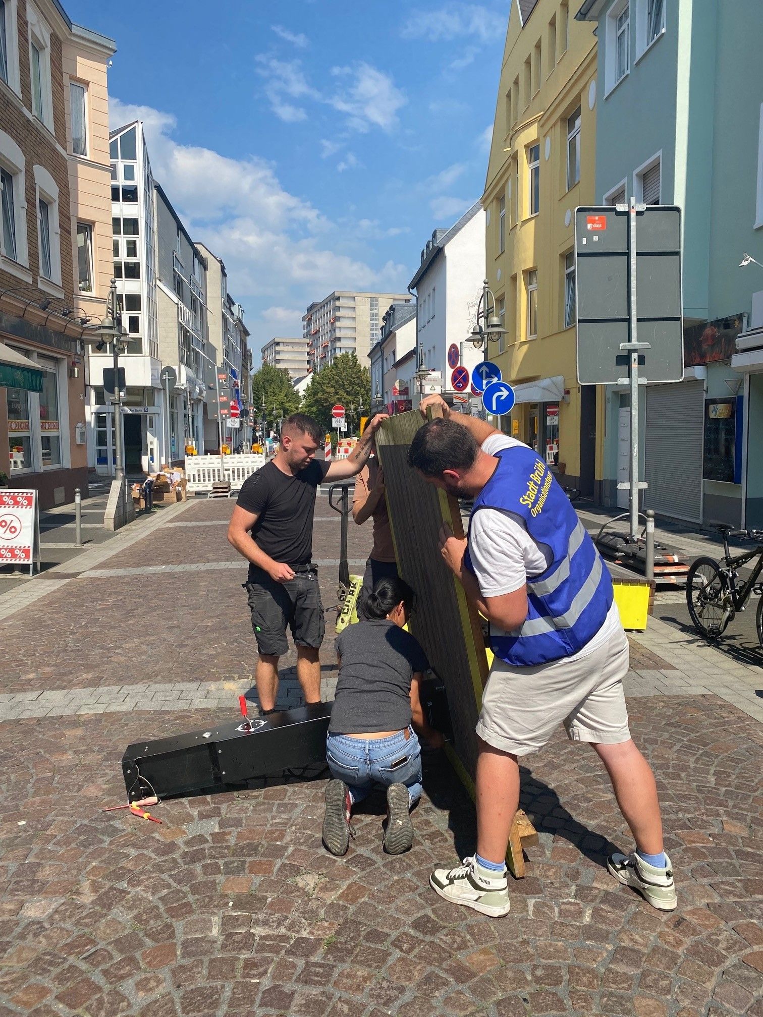 Das Bild zeigt Mitarbeiter der Stadt, die mit vereinten Kräften auf der Kölnstraße die Bodenplatte für eine Fahrradreparaturstation halten, um Schrauben anzubringen.