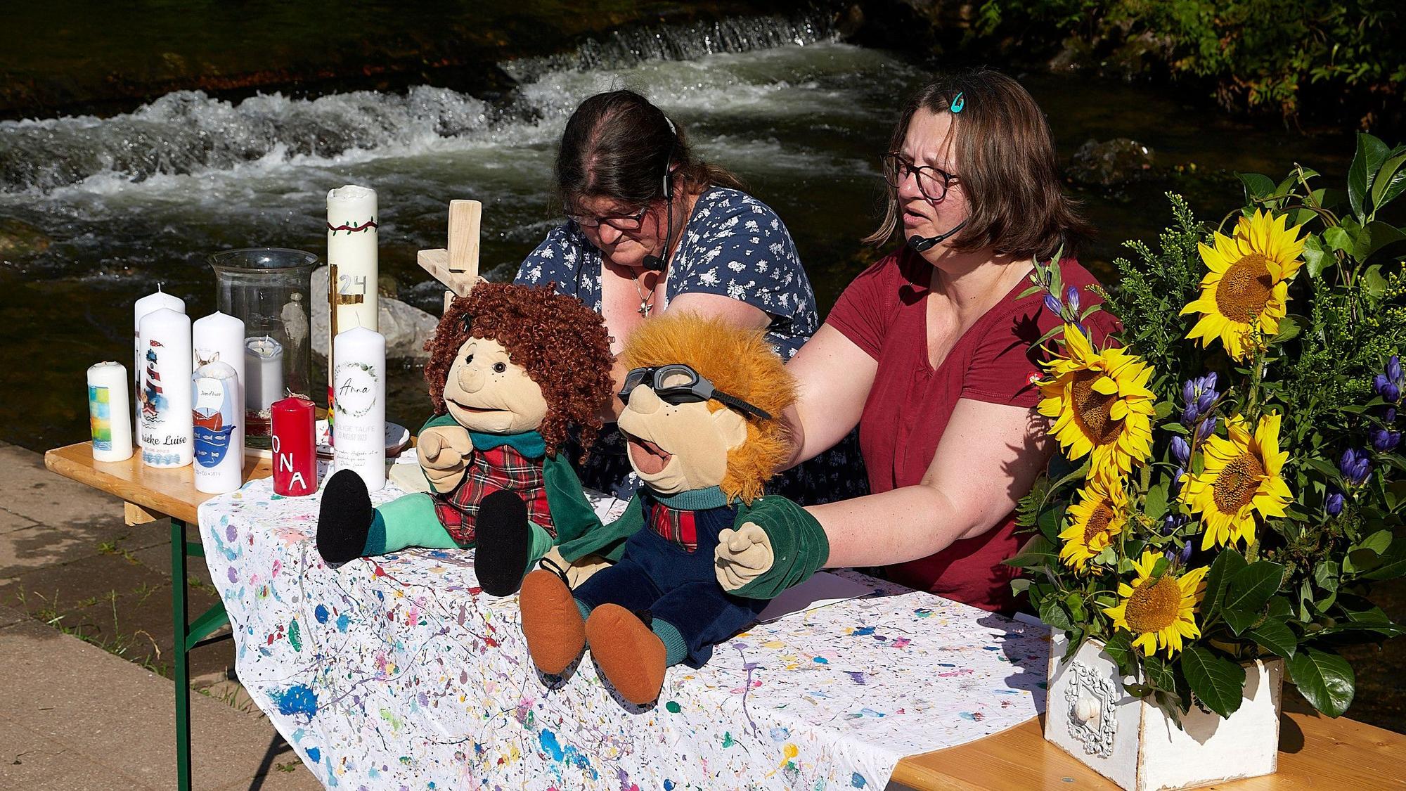 Zwei Frauen, Conny Friedrichs (l.) und Brigitte Vogel-Joswig, sitzen mit zwei Spielpuppen an einem Tisch. Der Tisch ist mit Kerzen und Blumen geschmückt.