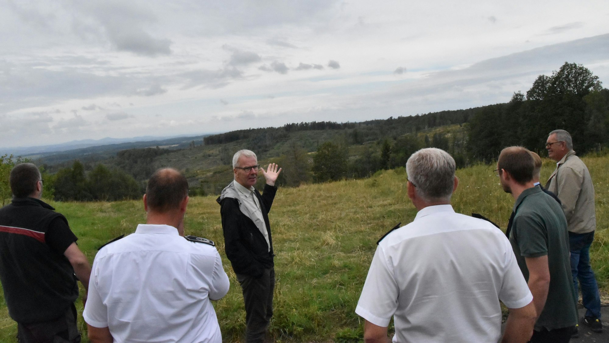 Mehrere Männer stehen auf einer Wiese, im Hintergrund ist ein Wald und der Höhenzug des Siebengebirges zu sehen.