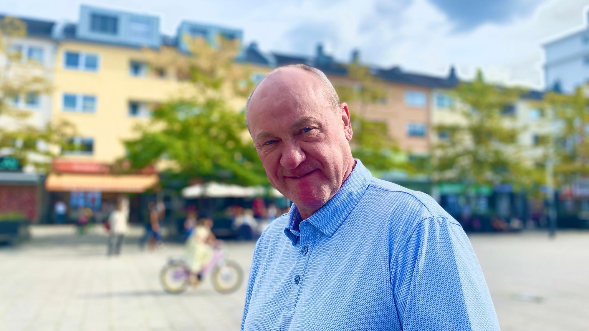 Ein mittelalter Mann mit wenigen Haaren und blauem Hemd steht auf einem Marktplatz.