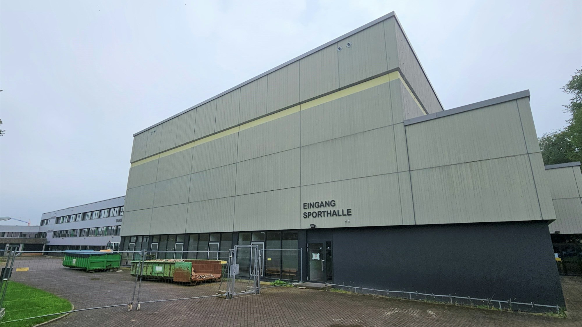 Das Bild zeigt die Sporthalle des Berufskollegs Eifel in Kall. Vor dem Gelände stehen Bauzäune und Container.