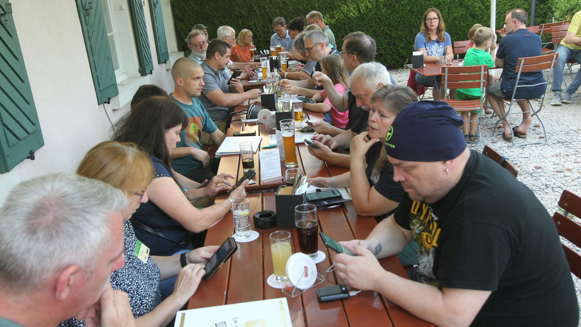 Beim Treffen der Fan-Community an den Holztischen in einem Biergartem sind alle mit ihren Handys beschäftigt.
