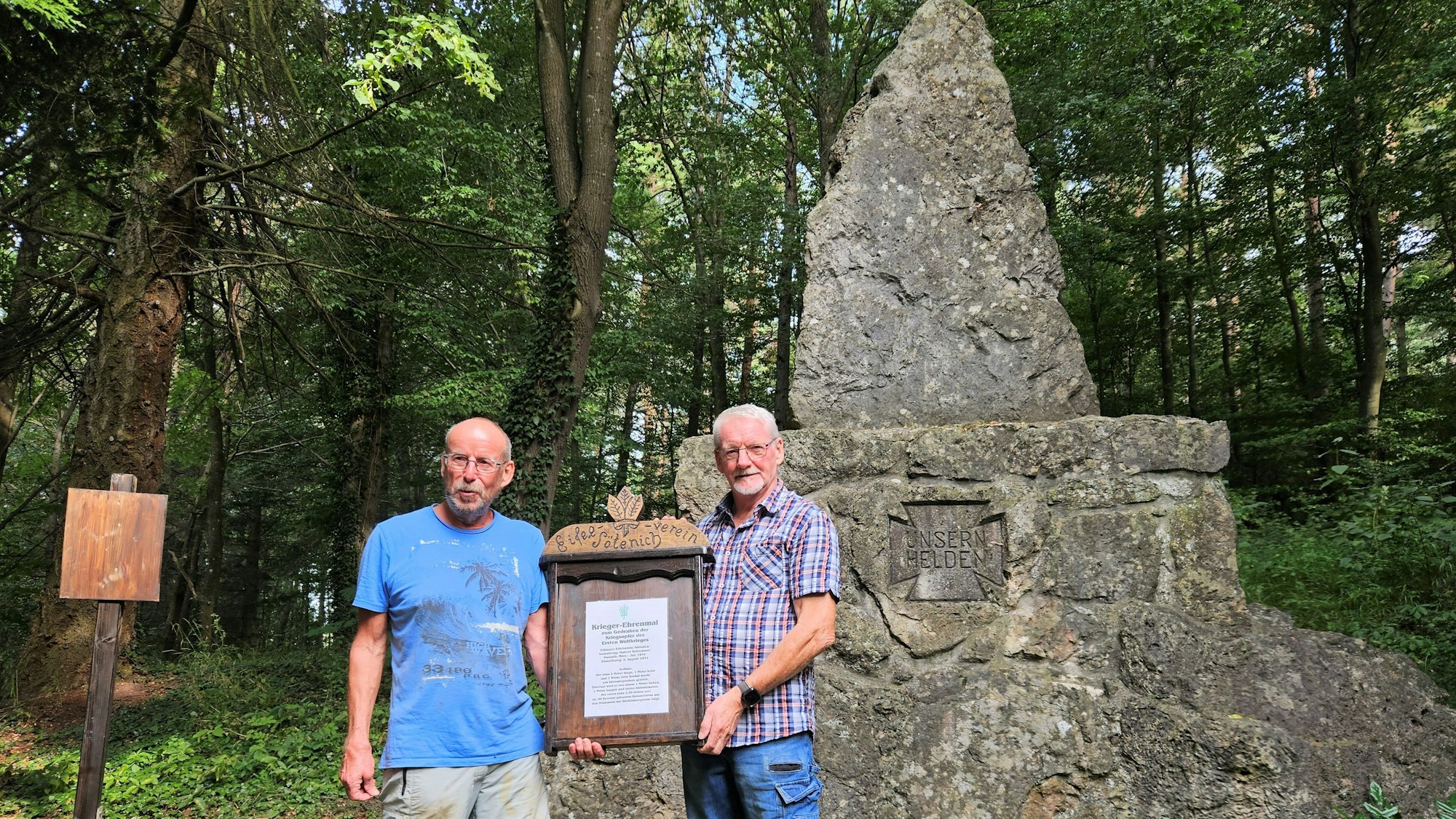 Wanderwart Karl-Heinz Geschwind (r.) und Wegewart Wilfried Schäfer halten eine Infotafel in den Händen. Sie stehen vor einem steinernen Ehrenmal.