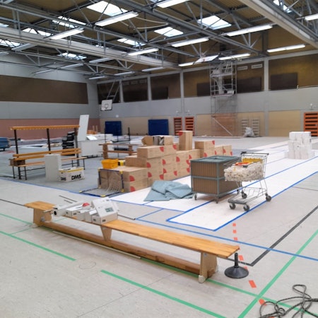 Das Bild zeigt eine Turnhalle. Auf einer Sportbank liegen Pakete. Große Kartons sind in der Mitte der Halle gelagert. Ein Einkaufswagen ist mit Plastik befüllt. An der Wand der Tundhalle steht ein Gerüst.