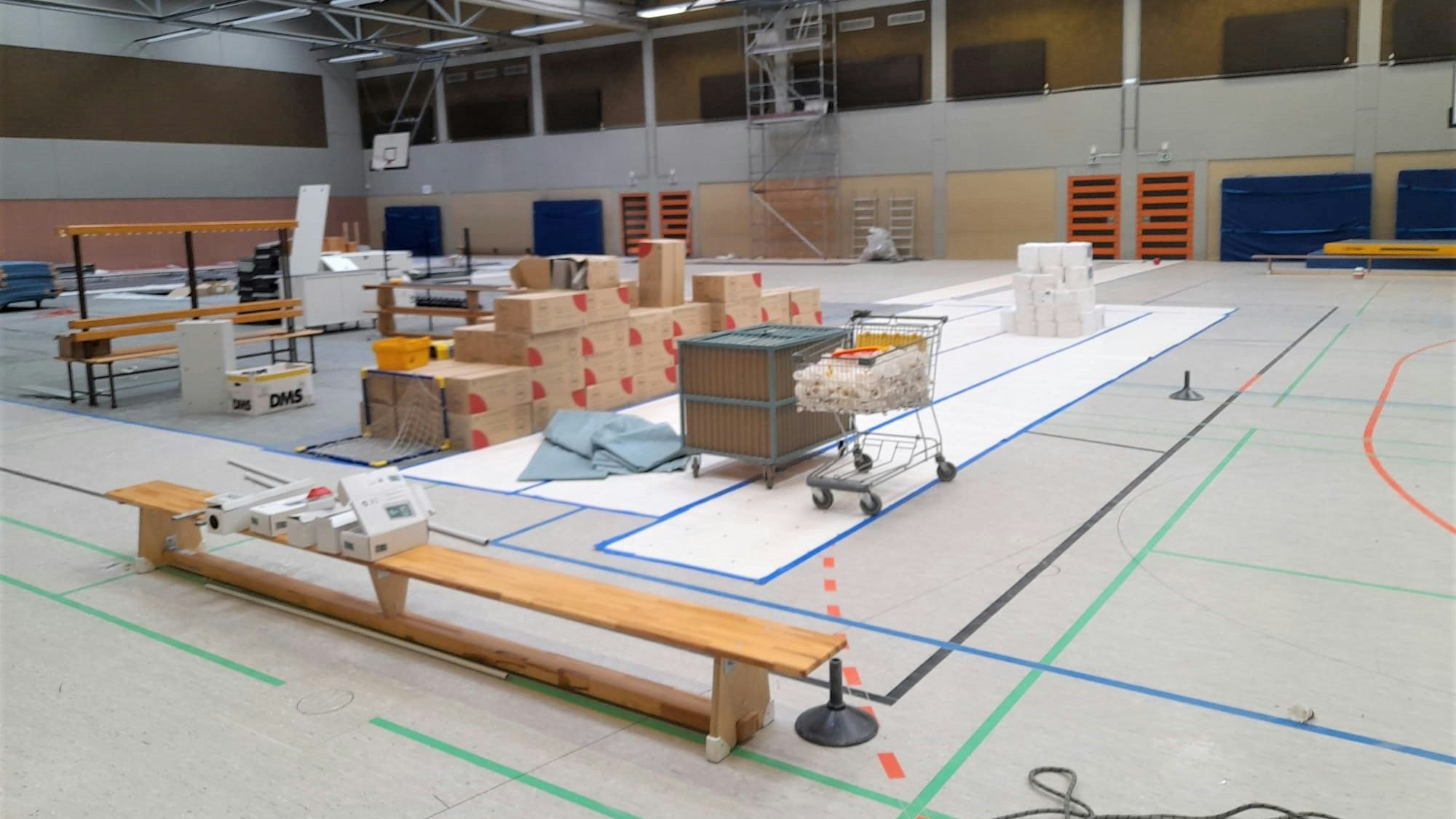 Das Bild zeigt eine Turnhalle. Auf einer Sportbank liegen Pakete. Große Kartons sind in der Mitte der Halle gelagert. Ein Einkaufswagen ist mit Plastik befüllt. An der Wand der Tundhalle steht ein Gerüst.