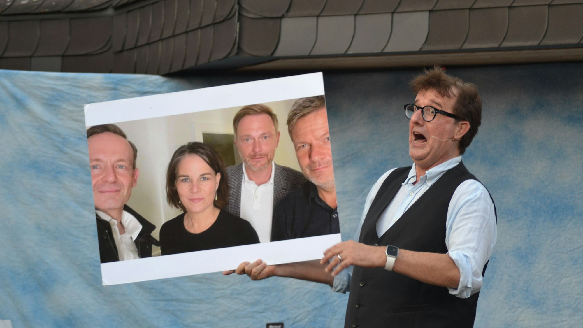 Ein Mann mit Brille und Weste hält ein Foto von Volker Wissing, Annalena Baerbock, Christian Lindner und Robert Habeck hoch und zieht eine Grimasse.