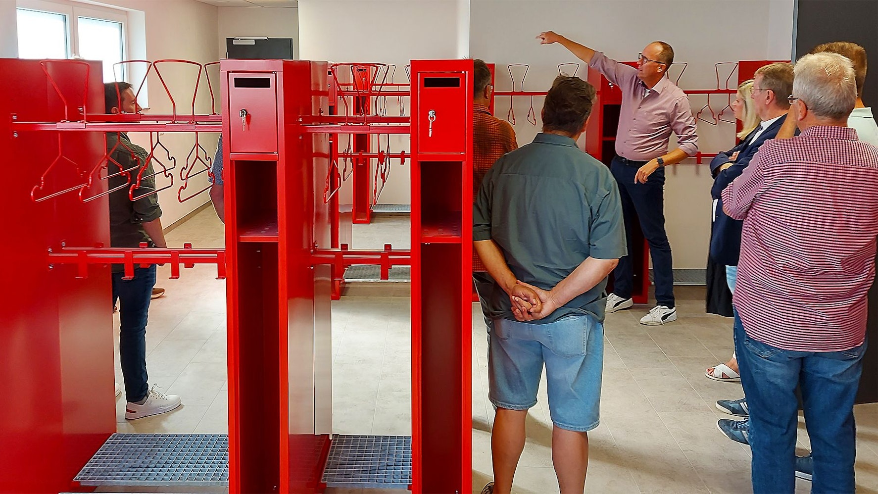 Im Umkleidebereich des Lommersdorfer Gerätehauses sind rote Spinde zu sehen. Rechts steht eine Gruppe Menschen, im Hintergrund ist Löschgruppen-Chef Torsten Luxen, der den anderen etwas erklärt.