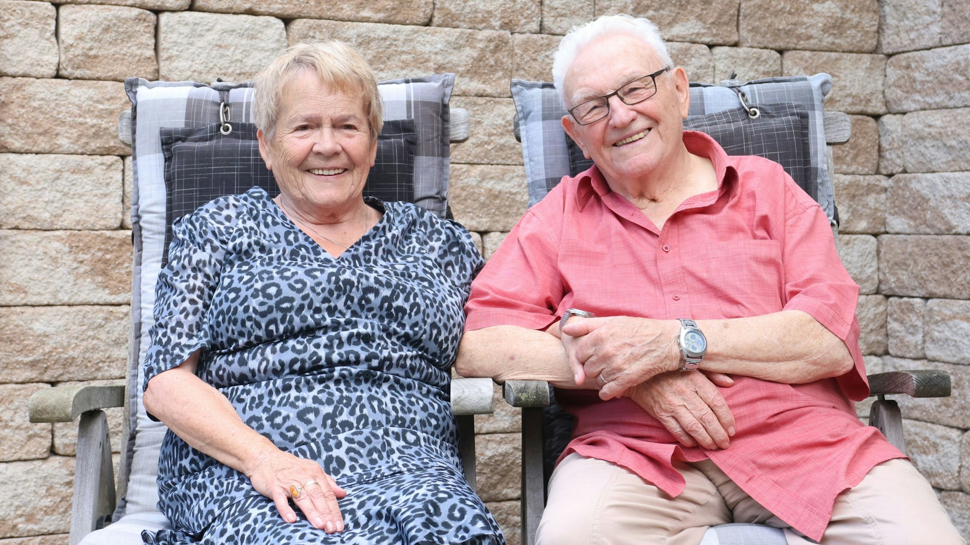 Ein älteres Ehepaar sitzt in Gartenstühlen nebeneinander. Erhält ihre Hand.