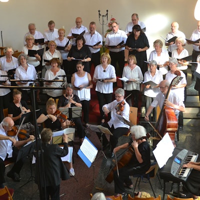 Das Bild zeigt einen Kirchenchor. Im Vordergrund sitzen Musiker mit Cello, Geigen, E-Piano und Flöten. Im Hintergrund stehen die Sänger und Sängerinnen.