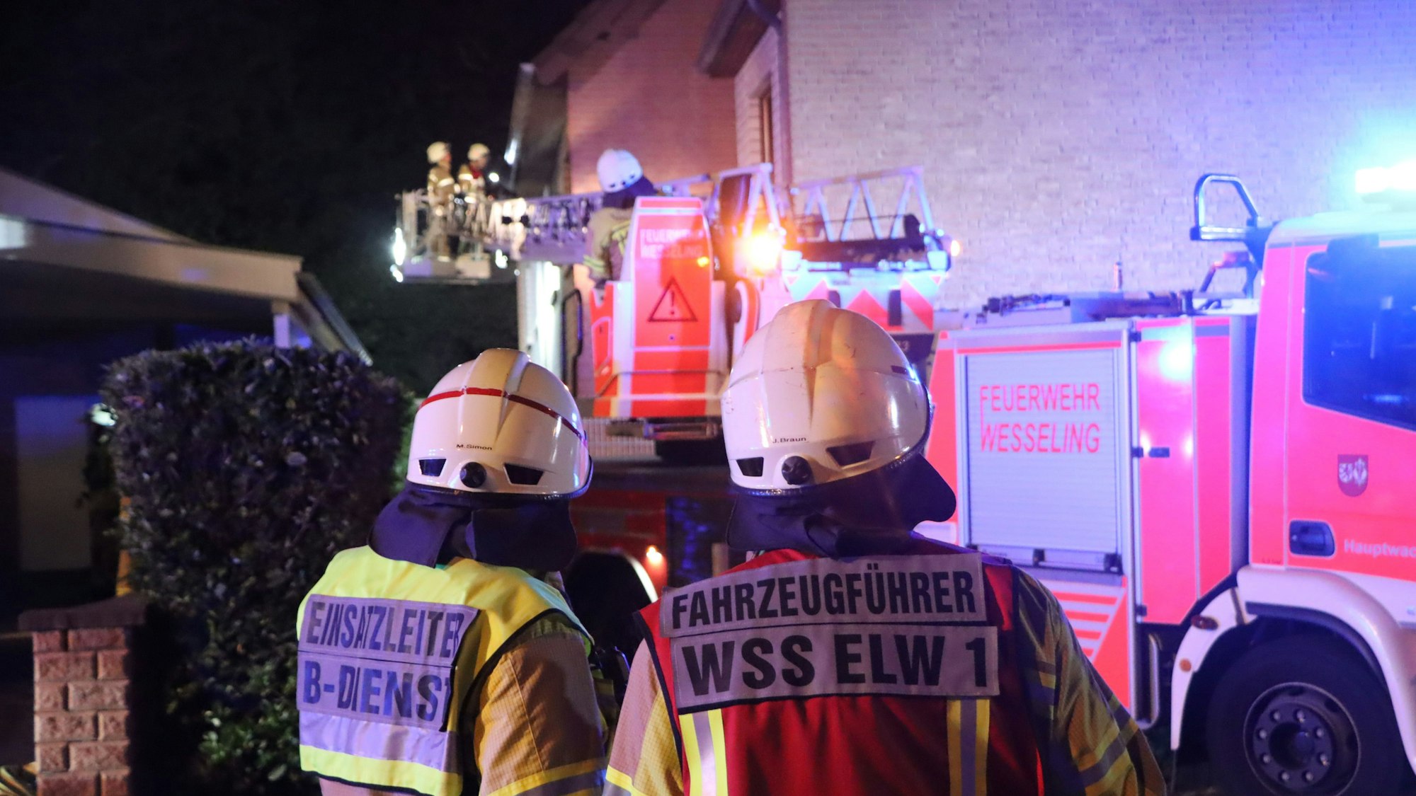 Zwei Feuerwehrleute stehen vor dem Appartementhaus. Im Hintergrund ist die Drehleiter mit zwei weiteren Einsatzkräften zu sehen.
