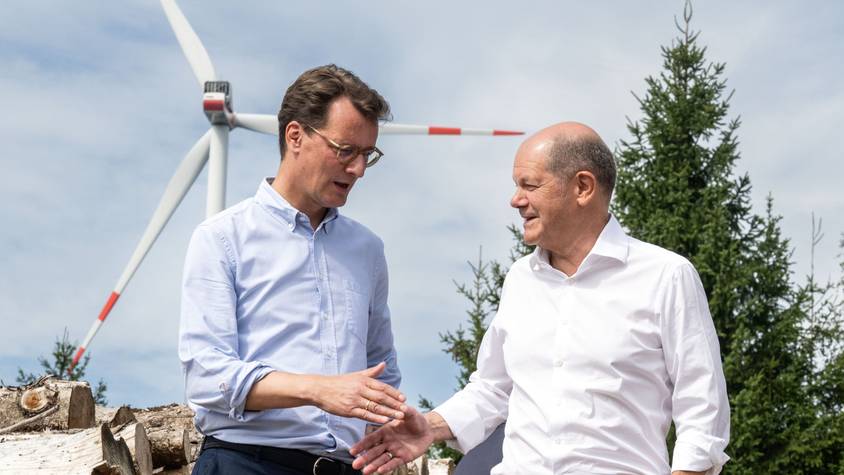 Bundeskanzler Olaf Scholz und Hendrik Wüst reichen sich die Hand. Im Hintergrund ist ein Windrad.