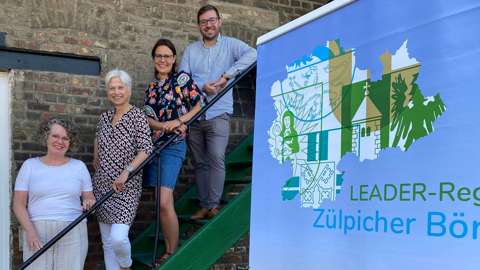 Auf einer grünen Treppe vor einer Wand stehen: die Weilerswister Bürgermeisterin Anna-Katharina Horst (v.l.), als Projektträgerin Marietta Thien, Projektmitarbeiterin Sarah Rodewald und Regionalmanager des LAG Zülpicher Börde, Sebastian Duif.