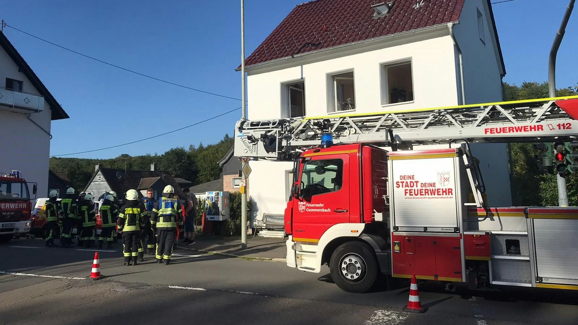 Vor einem Haus steht ein Einsatzfahrtzeug samt Drehleiter der Feuerwehr. Daneben stehen mehrere Feuerwehrleute.