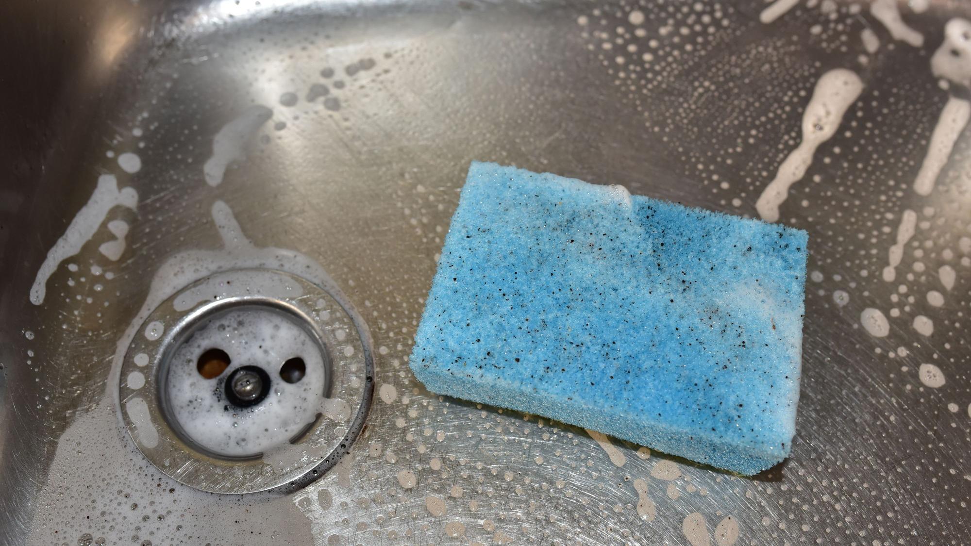 Ein dreckiger, hellblauer Spülschwamm liegt in einer Spüle, in der außerdem Rückstände von Spülmittel zu sehen sind.