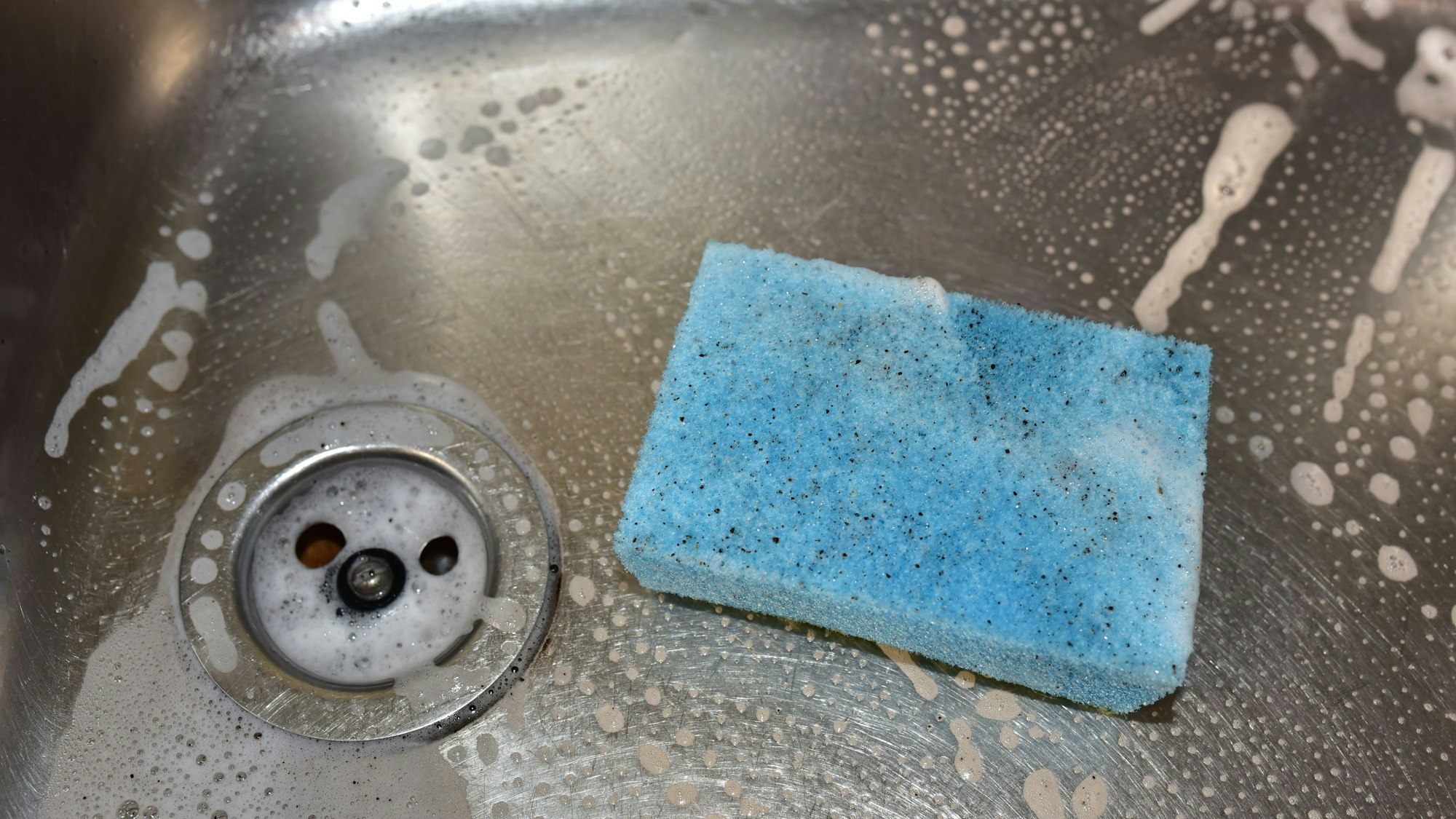 Ein dreckiger, hellblauer Spülschwamm liegt in einer Spüle, in der außerdem Rückstände von Spülmittel zu sehen sind.