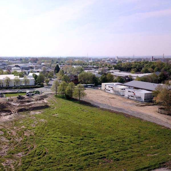 Aufsicht von oben auf ein gerodetes Areal mit zerfahrener Wiese, an deren Rand Baustellenfahrzeuge stehen. Bebauung ist sichtbar.