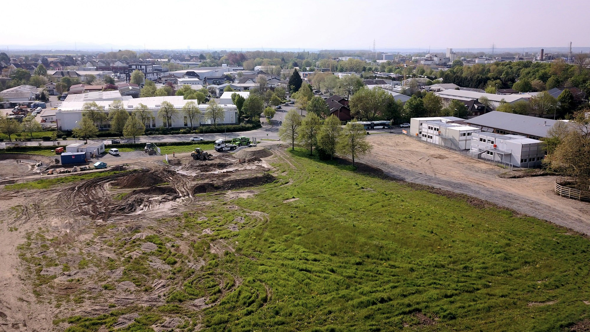 Aufsicht von oben auf ein gerodetes Areal mit zerfahrener Wiese, an deren Rand Baustellenfahrzeuge stehen. Bebauung ist sichtbar.
