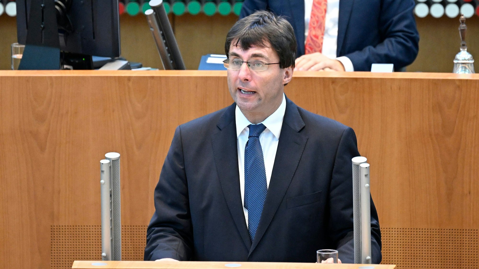 Marcus Optendrenk (CDU) spricht an einem Rednerpult - im Hintergrund ist eine hölzerne Abgrenzung sowie eine Person im Anzug zu sehen.