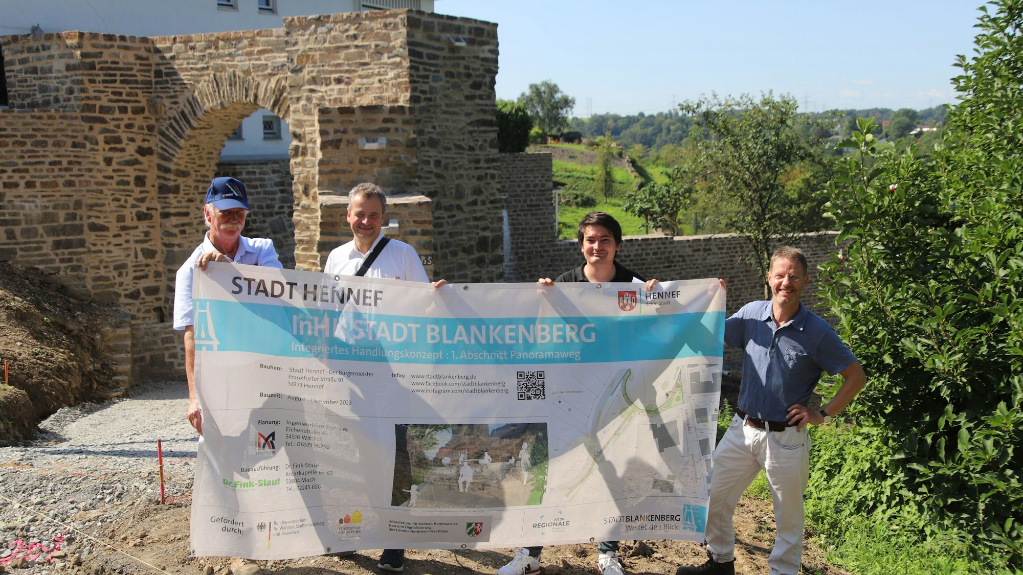 Vier Männer stehen vor einer alten Stadtmauer mit Durchgang und halten ein großes Planungsplakat.