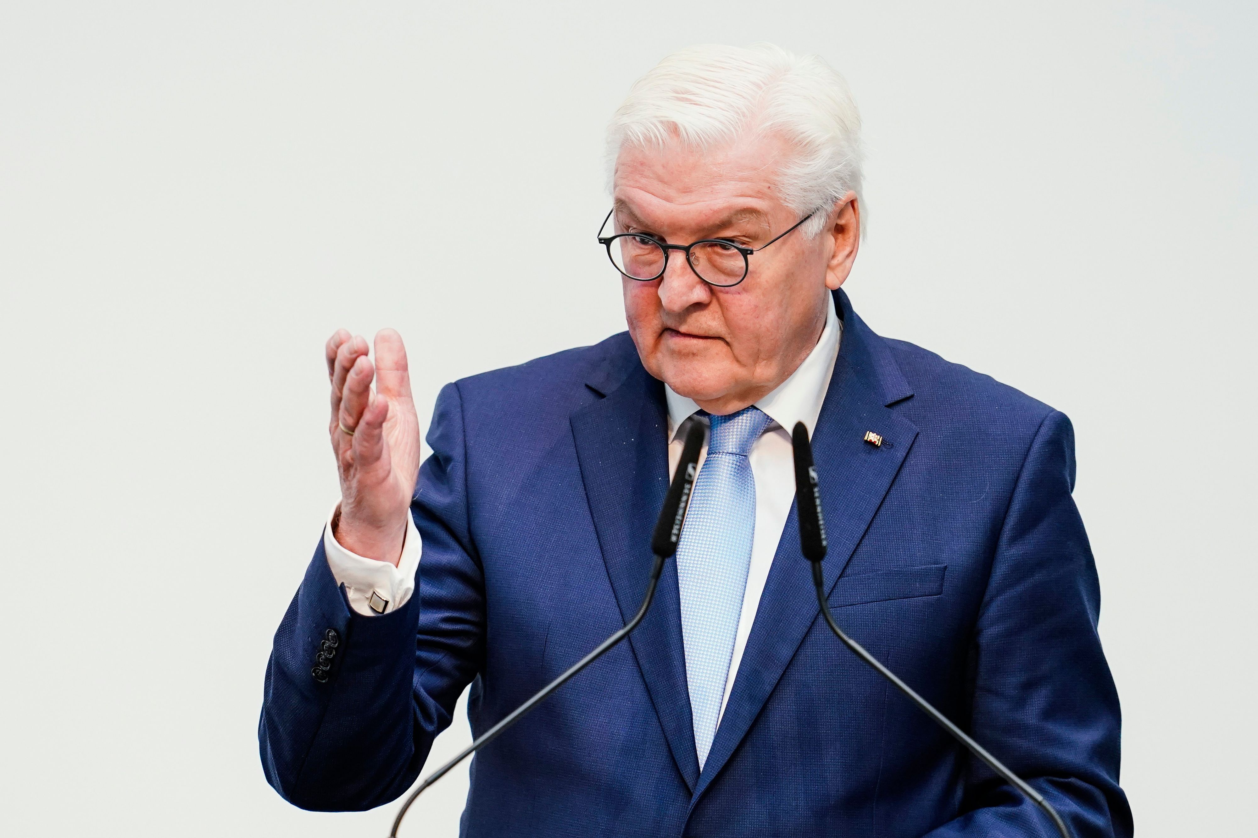 Bundespräsident Frank-Walter Steinmeier steht vor Mikrofonen und hebt eine Hand vor einem weißen Hintergrund.