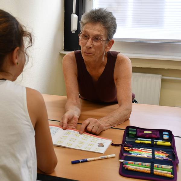 Ein Mädchen sitzt an der Schulbank vor einem Lehrbuch, dahinter eine Frau, die ihr hilft.