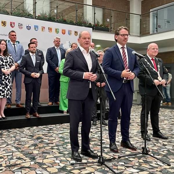Bundeskanzler Olaf Scholz steht mit zwei weiteren Männern vor mehreren Mikrofonen, im Hintergrund haben sich Bürgermeisterinnen und Bürgermeister aufgestellt.
