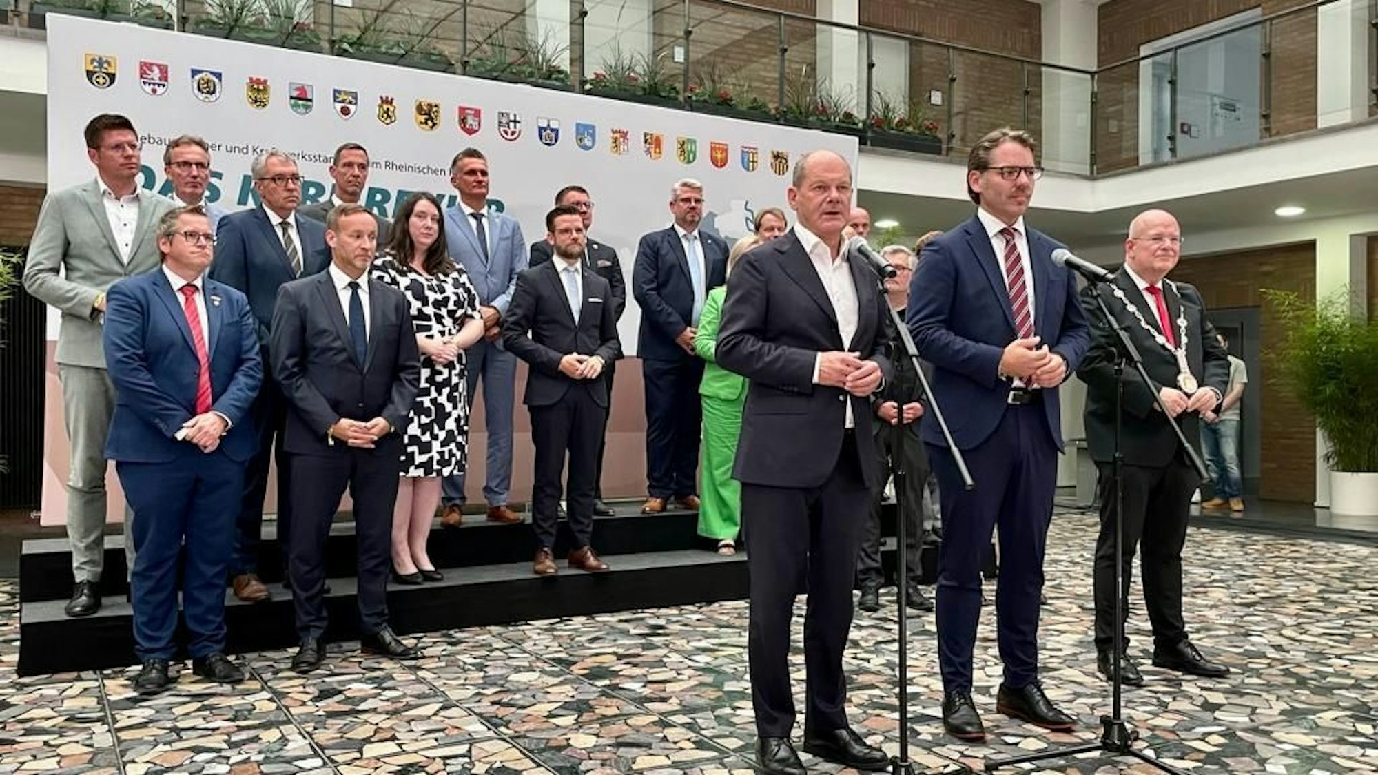 Bundeskanzler Olaf Scholz steht mit zwei weiteren Männern vor mehreren Mikrofonen, im Hintergrund haben sich Bürgermeisterinnen und Bürgermeister aufgestellt.