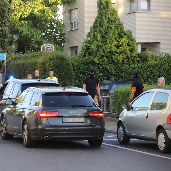 Mehrere Autos stehen auf einer Straße mit Mehrfamilienhäusern, Männer mit Sturmhauben stehen daneben.