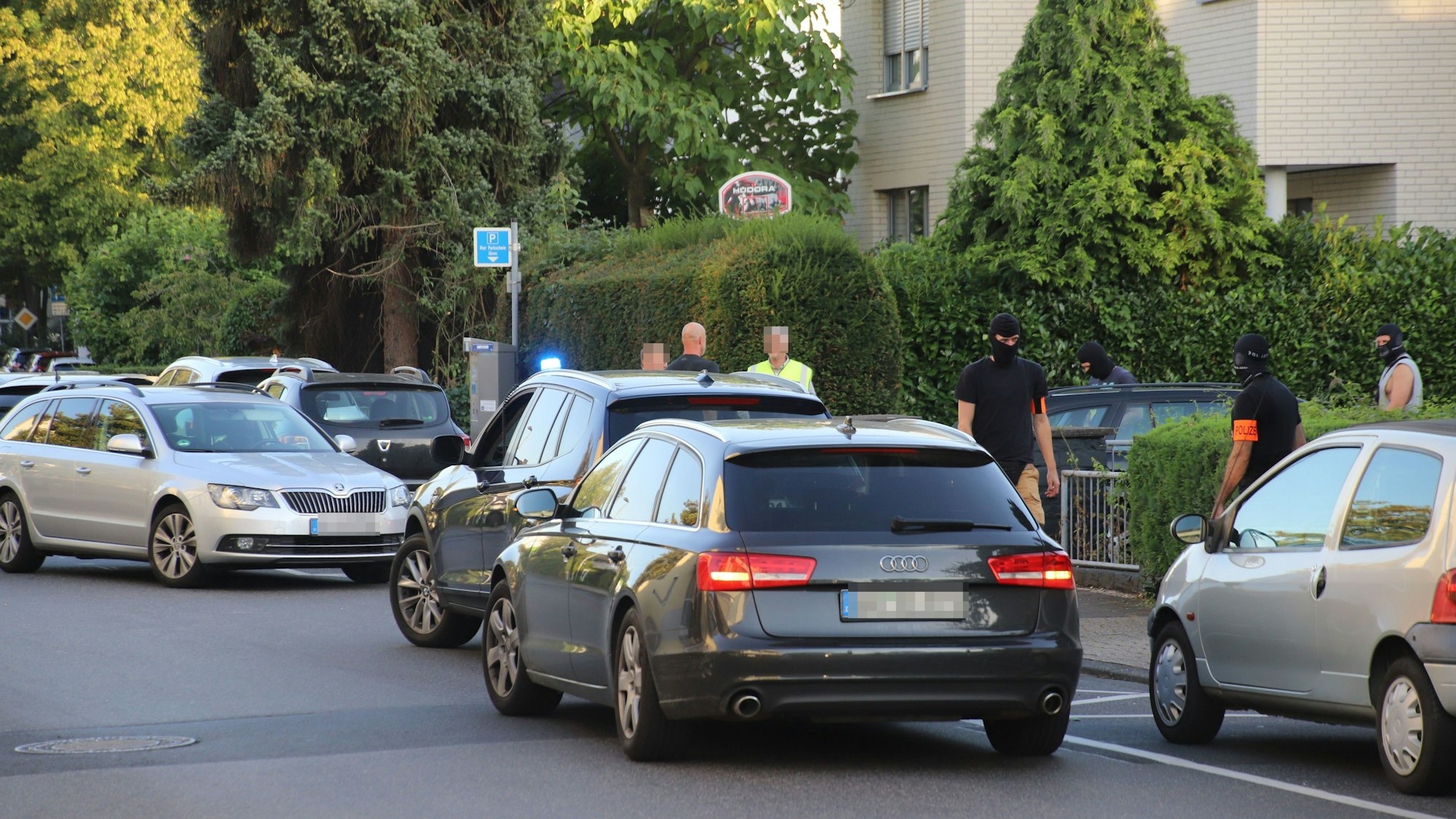 Mehrere Autos stehen auf einer Straße mit Mehrfamilienhäusern, Männer mit Sturmhauben stehen daneben.