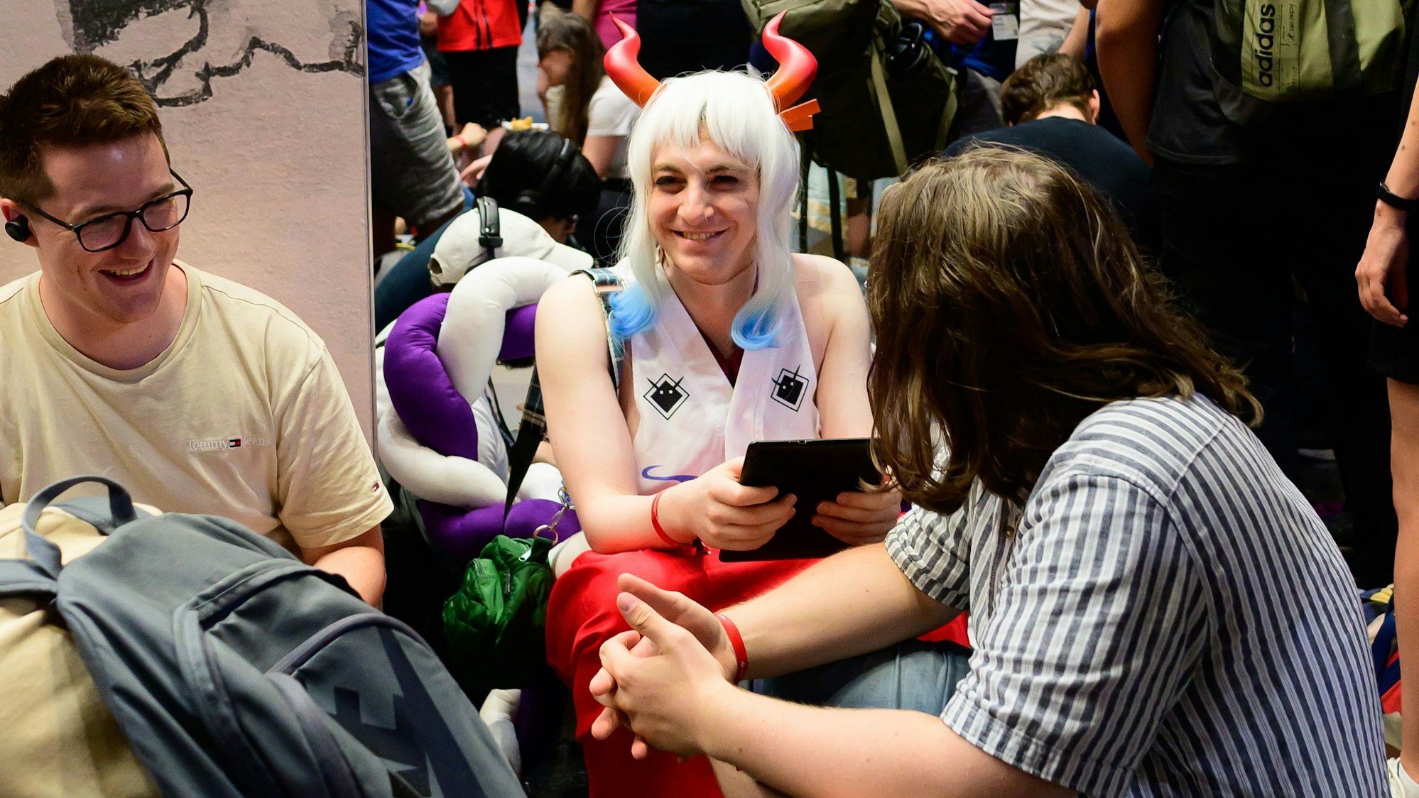 Drei Männer, einer von ihnen bunt kostümiert, sitzen in einer Warteschlange.