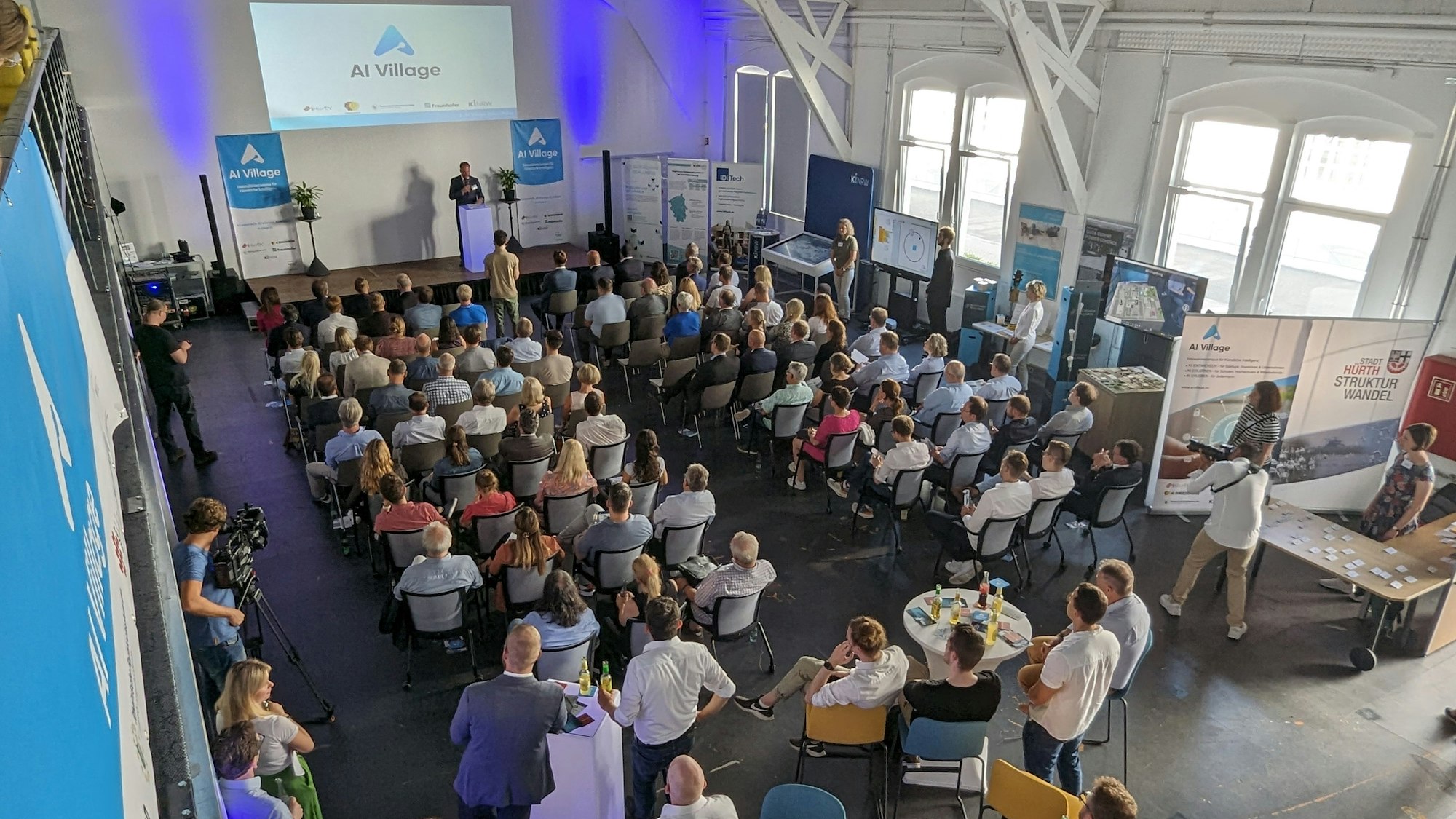 Volle Sitzreihen und Stellwände in einem Saal.