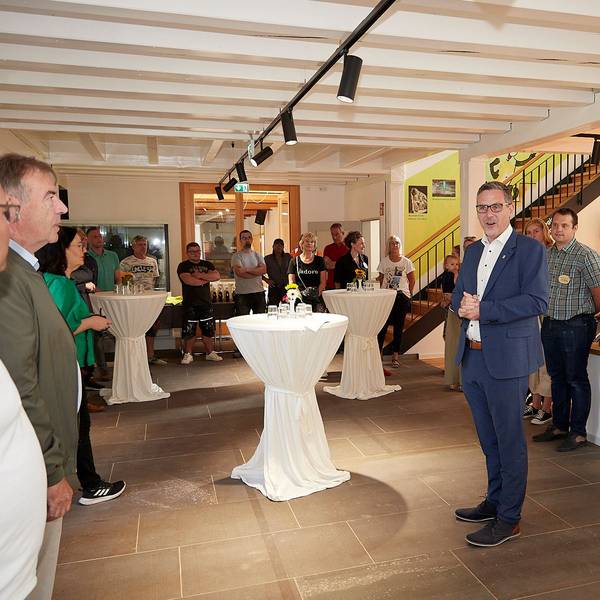 In einem großen Raum stehen Stehtische mit weißen Hussen. In der Mitte steht Nettersheims Bürgermeister Norbert Crump, an den Wänden zahlreiche andere Menschen. Sie feiern die Wiedereröffnung des Naturzentrums.