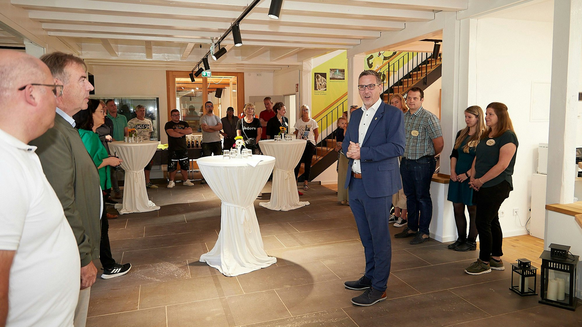 In einem großen Raum stehen Stehtische mit weißen Hussen. In der Mitte steht Nettersheims Bürgermeister Norbert Crump, an den Wänden zahlreiche andere Menschen. Sie feiern die Wiedereröffnung des Naturzentrums.