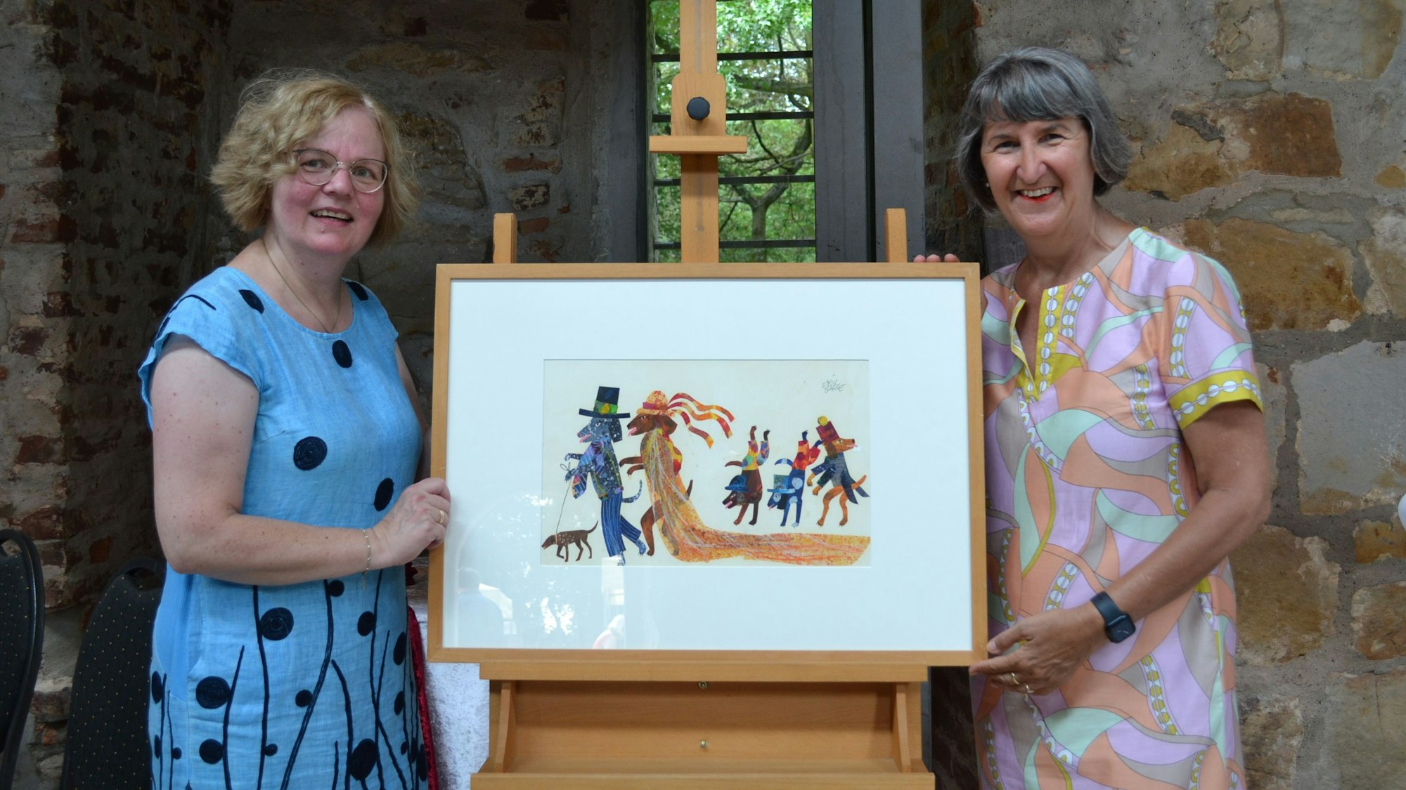 Zwei Frauen stehen rechts und links von einem gerahmten Bild, das eine bunte Zeichnung zeigt. Zwei Hunde in Zylinder und Schleier, hinter ihnen drei kleinere Hunde, alle tragen Hüte.