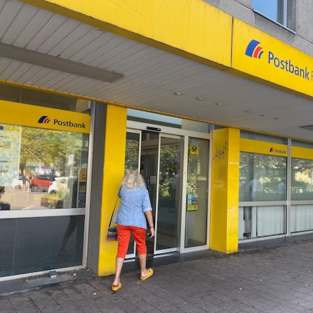 Eine Frau, die von hinten zu sehen ist, geht in die Filiale einer Postbank.
